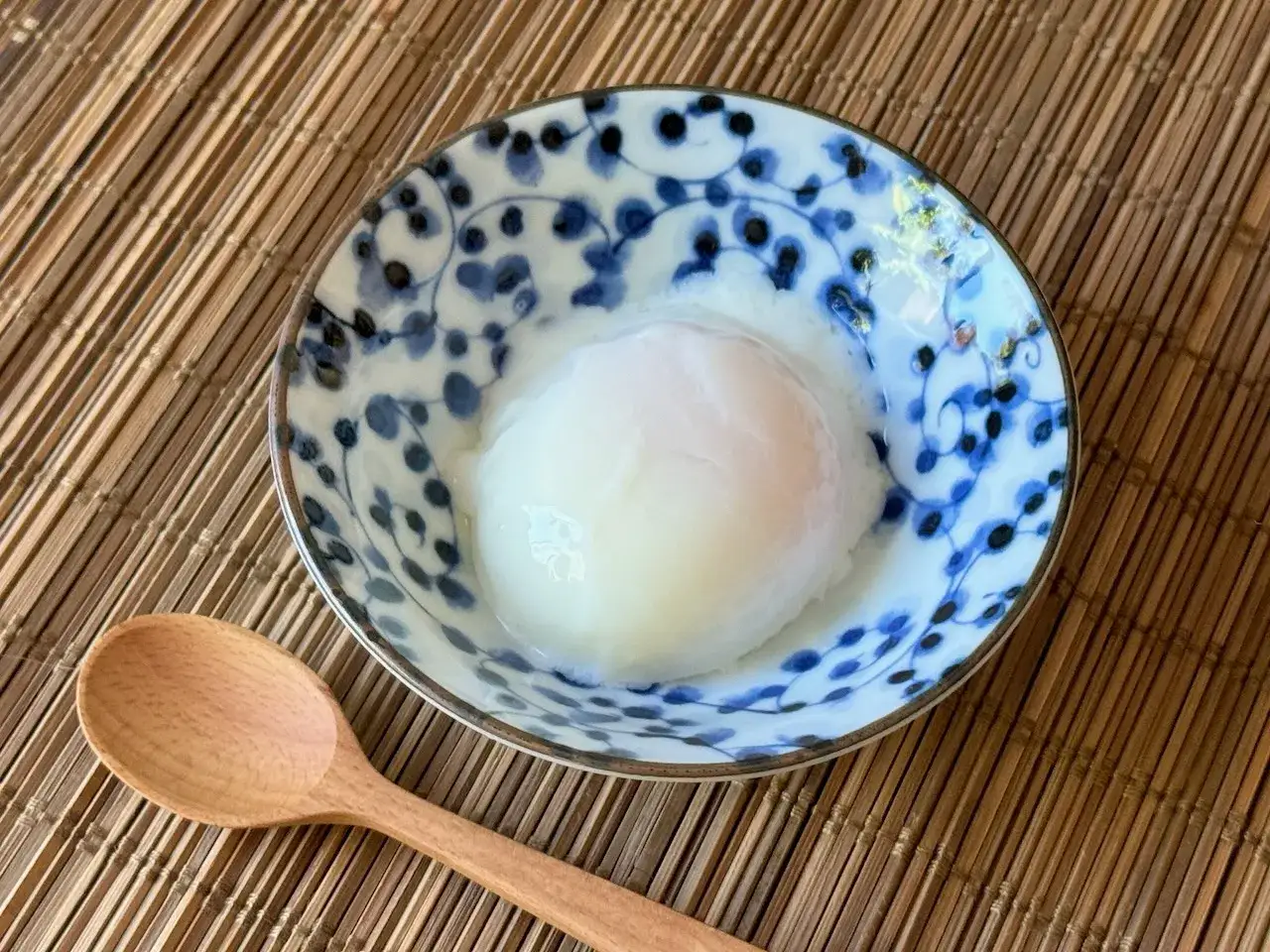A perfectly poached egg, a staple of Japanese cuisine, sits in a delicate blue and white bowl. A wooden spoon rests beside it, ready to scoop up this delicious breakfast.