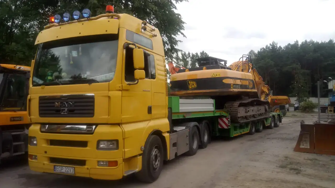 Żółta ciężarówka z naczepą do transportu maszyn budowlanych przewozi koparkę JCB.