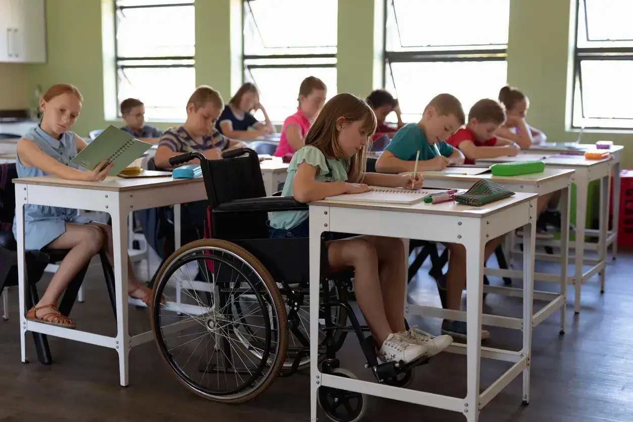Dzieci w klasie, jedna dziewczynka na wózku inwalidzkim, pisze w zeszycie. Kiedy złożyć wniosek o wydłużenie etapu edukacyjnego?