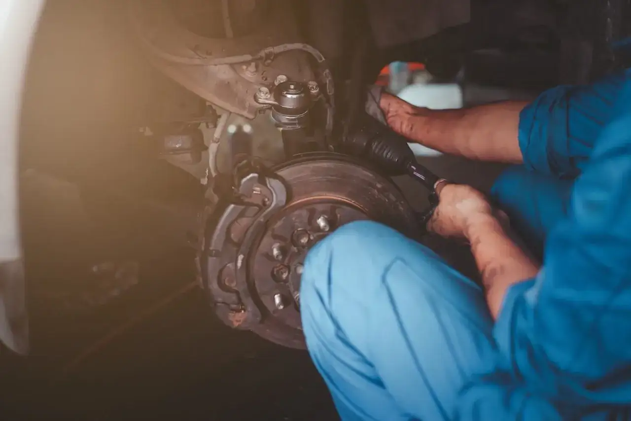 Mechanik w niebieskim kombinezonie pracuje przy kole samochodu, wykonując wymianę piasty.