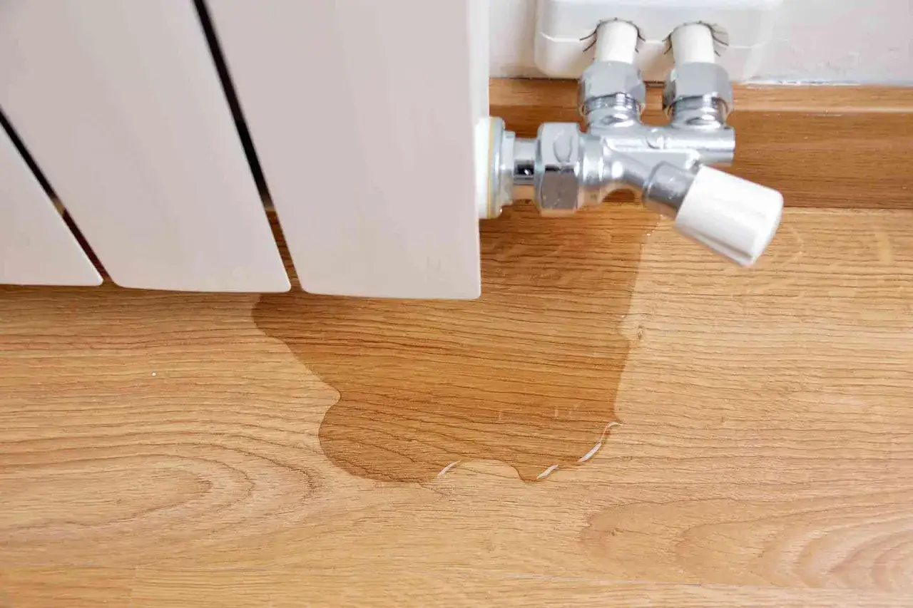 Un radiador que gotea sobre un suelo de madera. El agua forma un charco que se extiende.