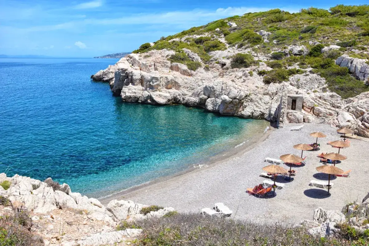 Rodos: turkusowe morze obmywa kamienistą plażę z leżakami i parasolami. Skaliste wybrzeże porośnięte zielenią.