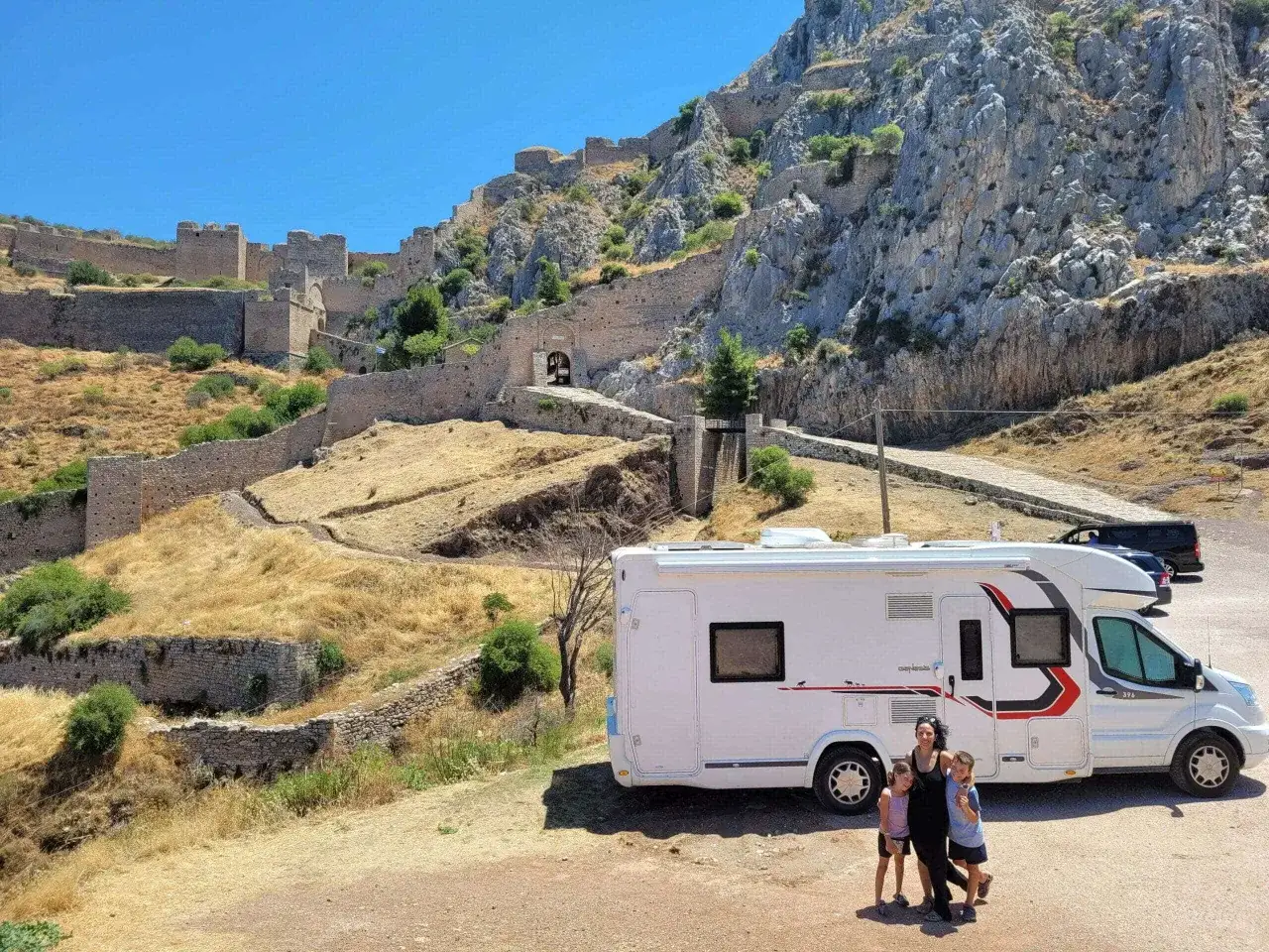 Une famille profite de son camping-car en Grèce, devant les ruines d'un ancien château sur une colline rocheuse.