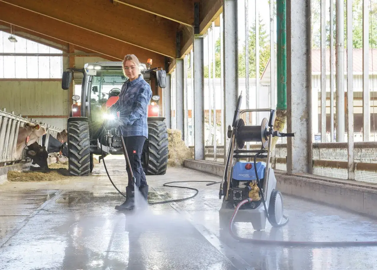 Frau reinigt Stallboden mit einem benzin hochdruckreiniger. Traktor und Kühe im Hintergrund.