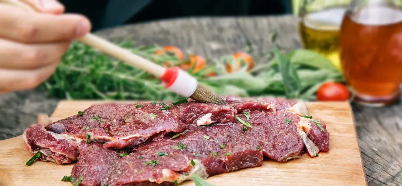 Fleisch marinieren wie lange? Ein Pinsel verteilt Marinade auf rohem Fleisch, Kr&auml;uter und Gew&uuml;rze sind bereits aufgetragen.