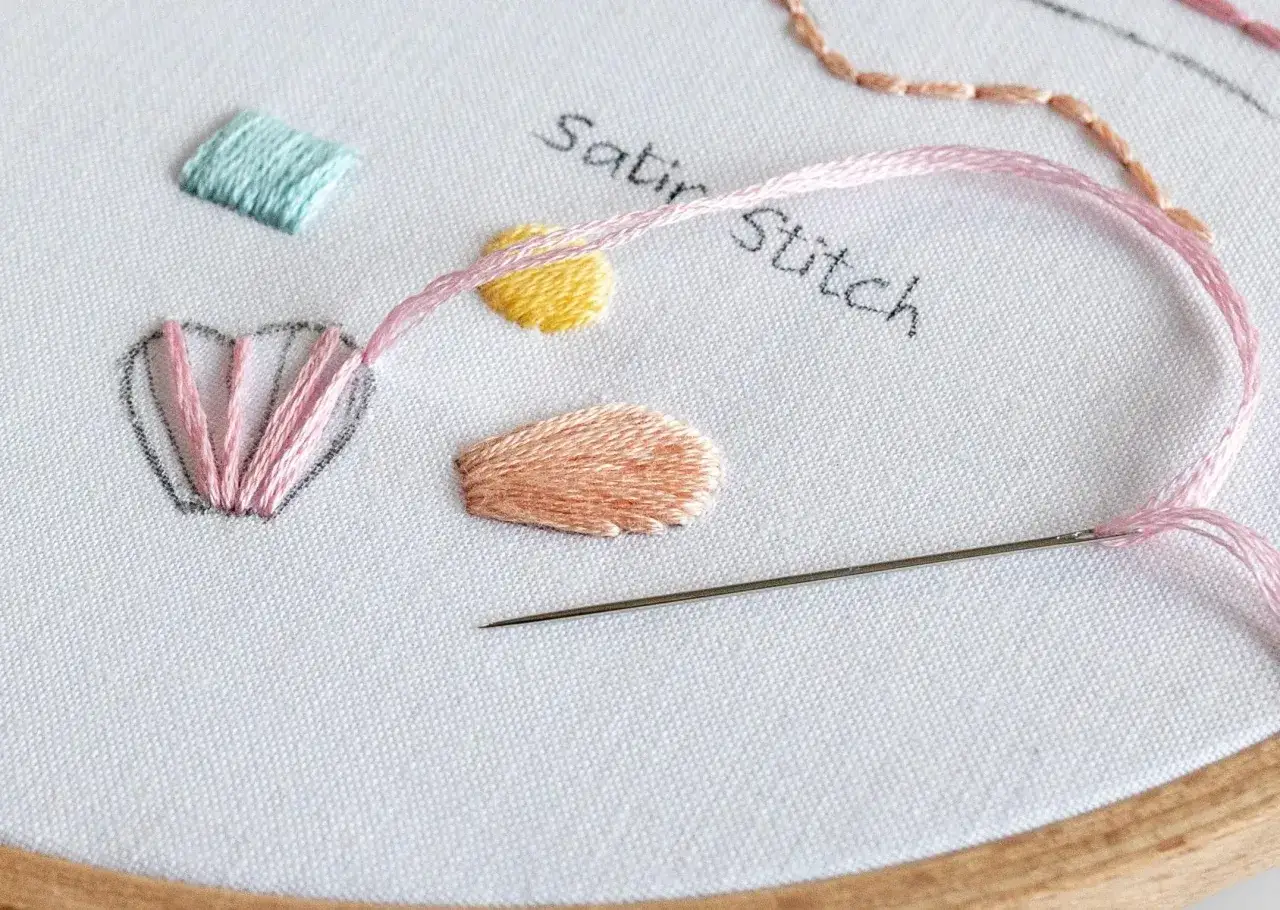 Bordado de muestra con hilo rosa y aguja. Se aprecian varios puntos, incluyendo el **satin stitch** en forma de pétalos y cuadrados.