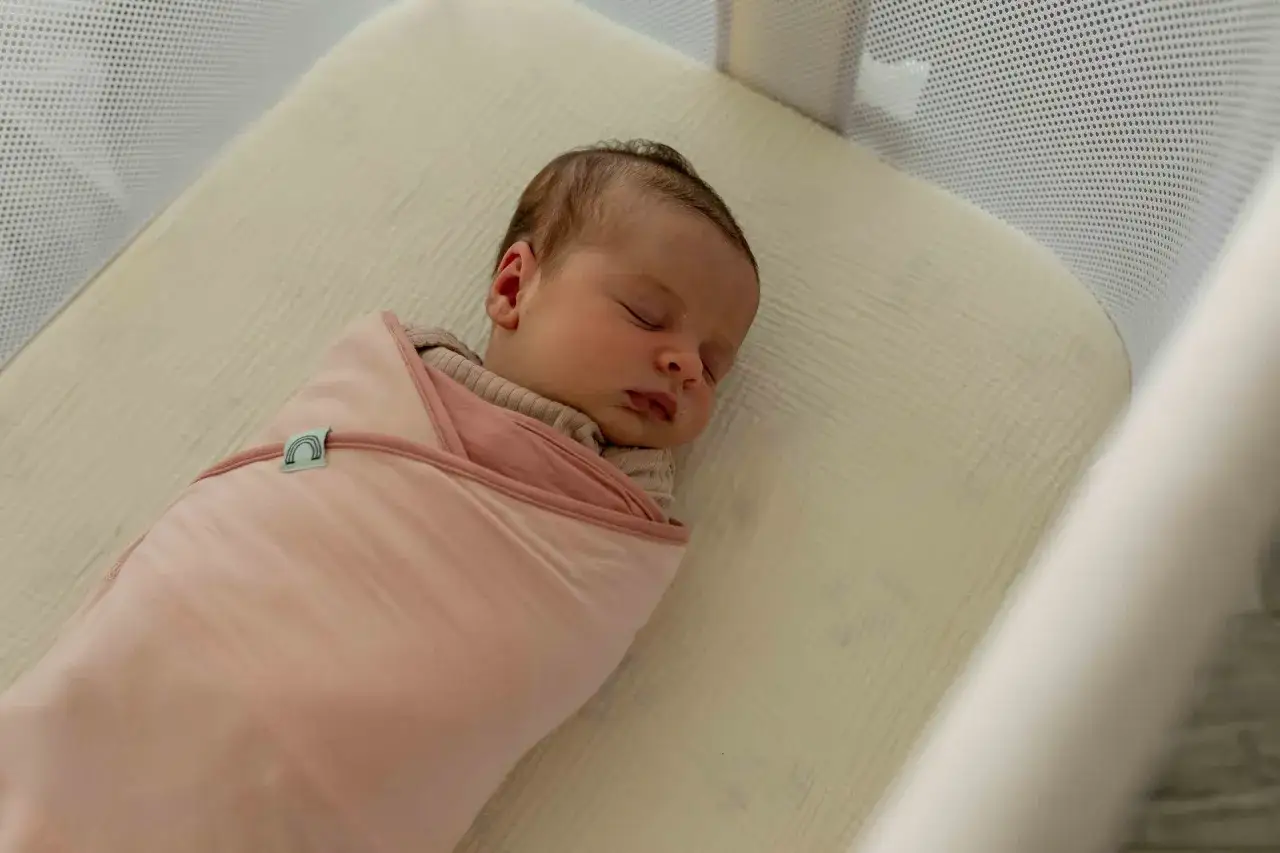 Śpiący 3 tygodniowy noworodek otulony w różowy kocyk z tęczą.