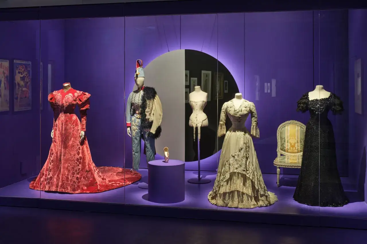 A museum display features historical fashion, including a red floral gown, a military uniform, a corset, and a black sequined dress, possibly inspired by Mathilde Favier's era.