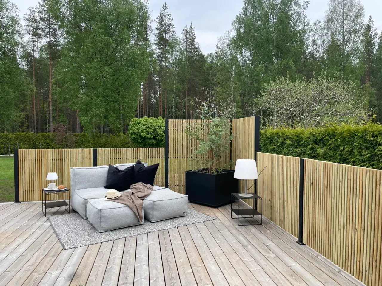 En mysig uteplats med grå soffa, kuddar och filt. Runtom ett modernt ribbstaket som ger insynsskydd mot skogen.