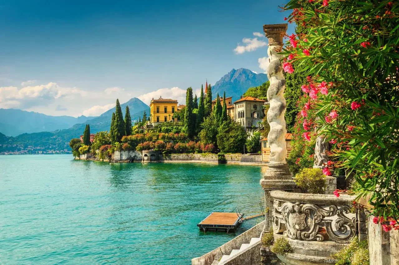 Vue d'un palais sur les lacs italiens, avec des montagnes en arrière-plan et des fleurs roses éclatantes au premier plan.
