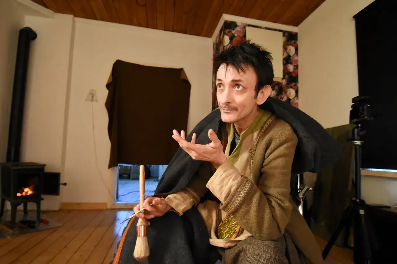 Andrea Lennartsson, med pensel och sax i hand, sitter vid en kamin och ber&auml;ttar.
