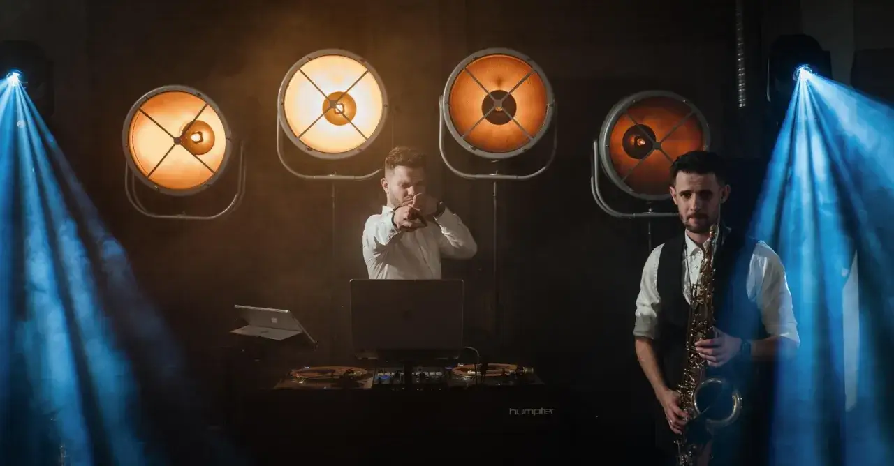 DJ i saksofonista tworzą idealną playlistę na wesele. W tle ciepłe światła i niebieskie snopy.