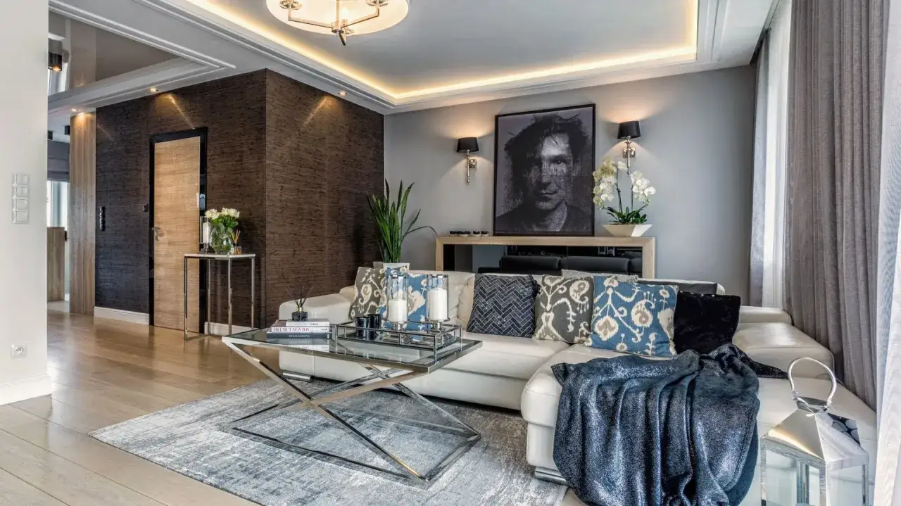 Eleganckie wnętrza modern classic: biała sofa, szare ściany, ozdobne poduszki, szklany stolik i portret na ścianie tworzą stylową przestrzeń.