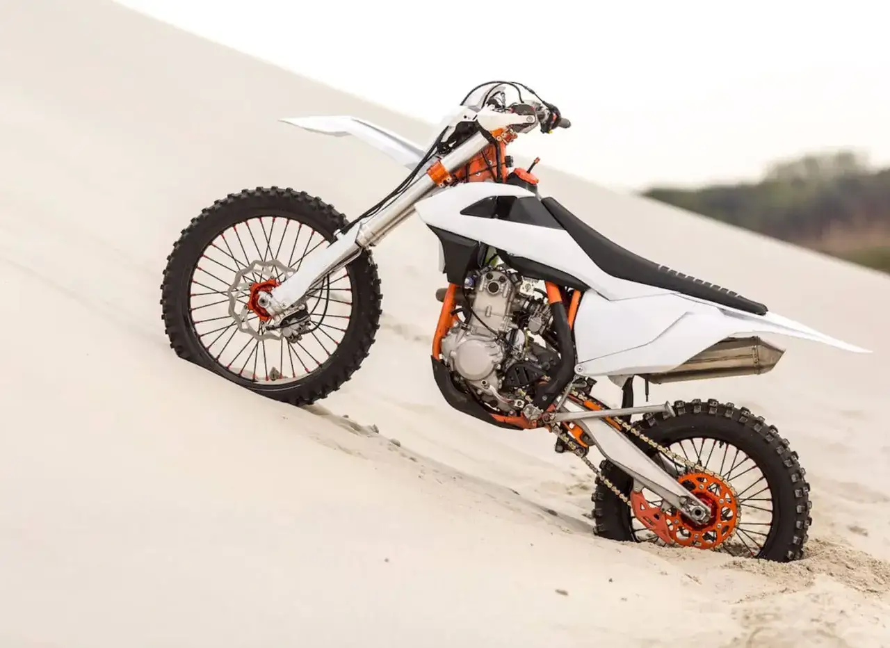 Una moto supermotard blanca y naranja se hunde en la arena, lista para la aventura.
