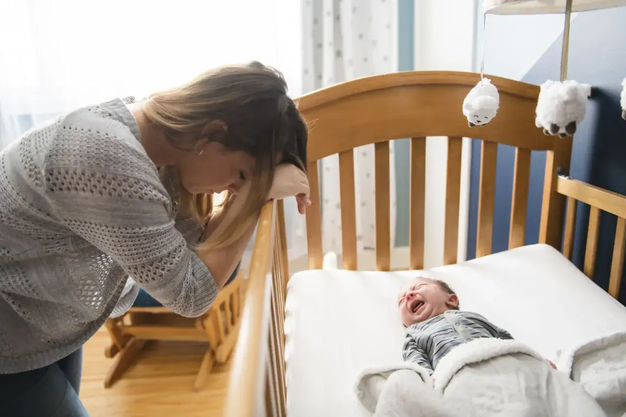 Zmęczona mama pochyla się nad ł&oacute;żeczkiem, gdzie płacze niemowlę. To trudny sen niemowlaka.