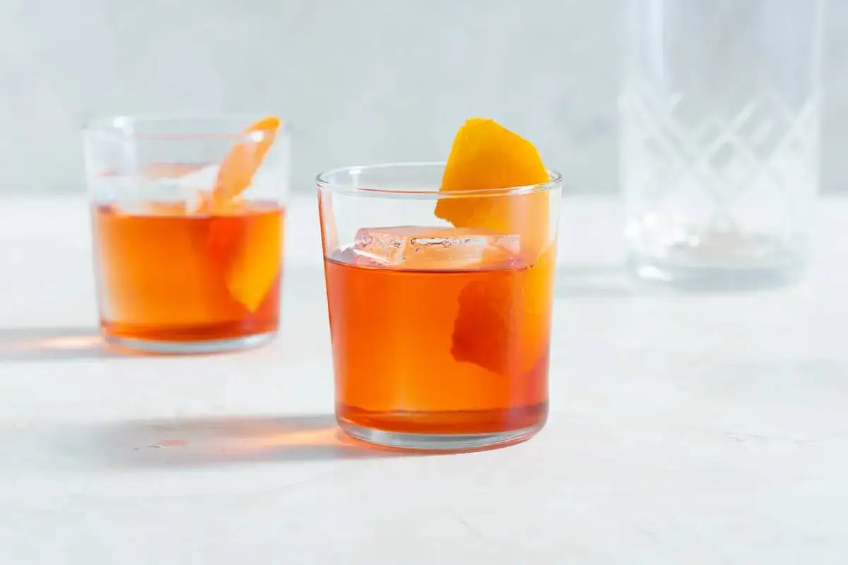Zwei leuchtend orangefarbene Cocktails, ein Negroni, mit Eis und Orangenschale garniert, stehen auf einem hellen Tisch.