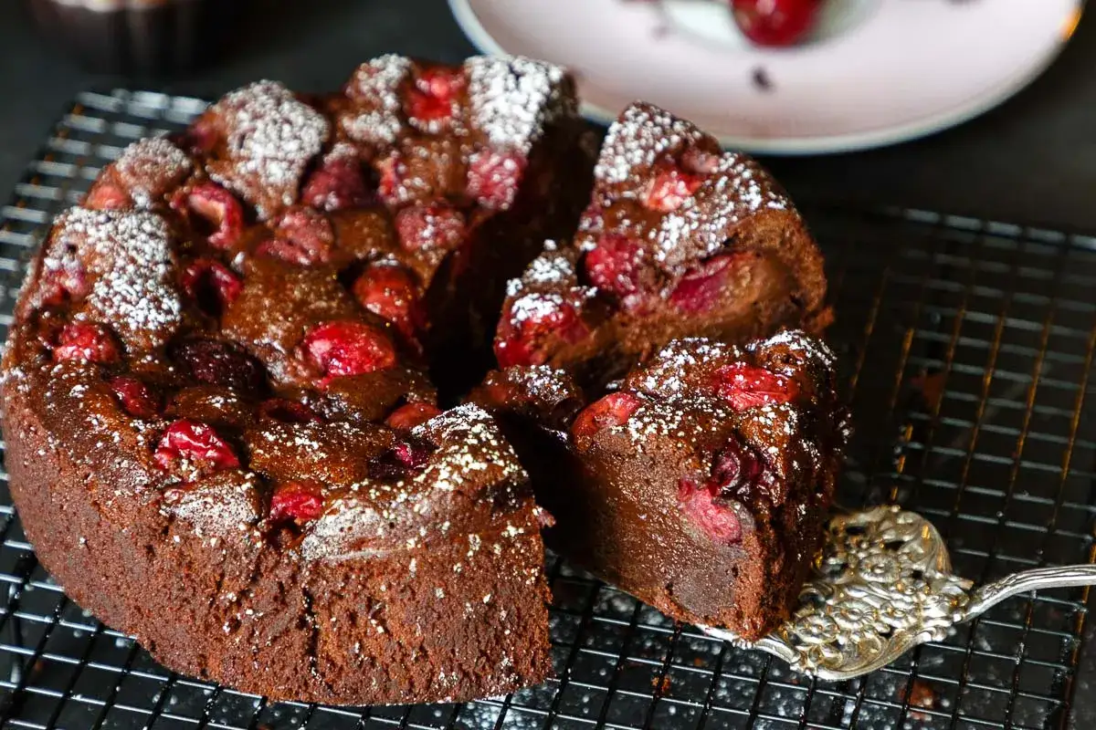 Saftiger Schoko-Kirsch-Kuchen aus der Springform, mit Puderzucker bestäubt und Kirschen garniert. Ein Stück wird gerade mit einer Tortengabel herausgehoben.