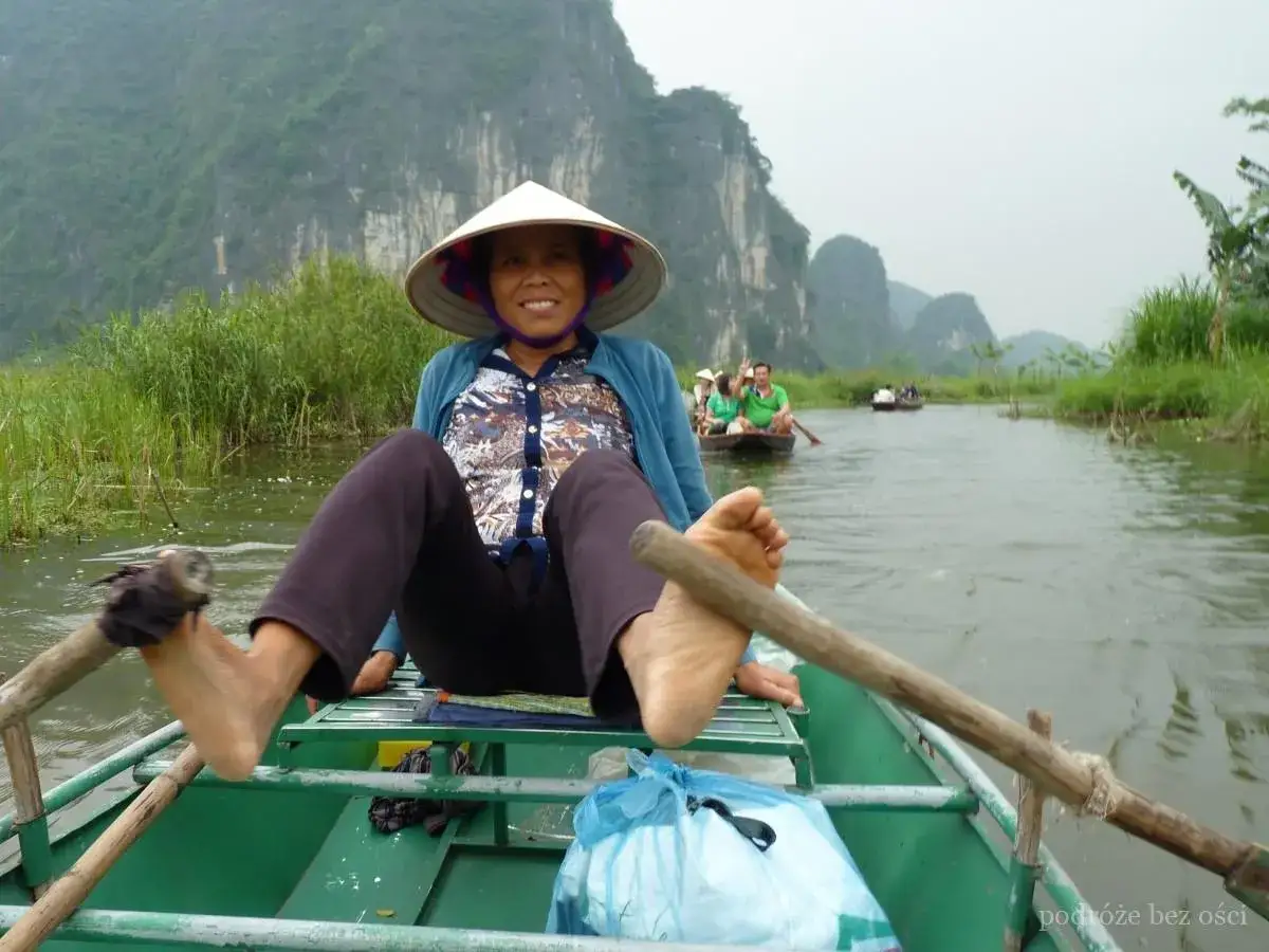 Kobieta w tradycyjnym kapeluszu wiosłuje łodzią po rzece w Ninh Binh, otoczona bujną zielenią i wapiennymi klifami.