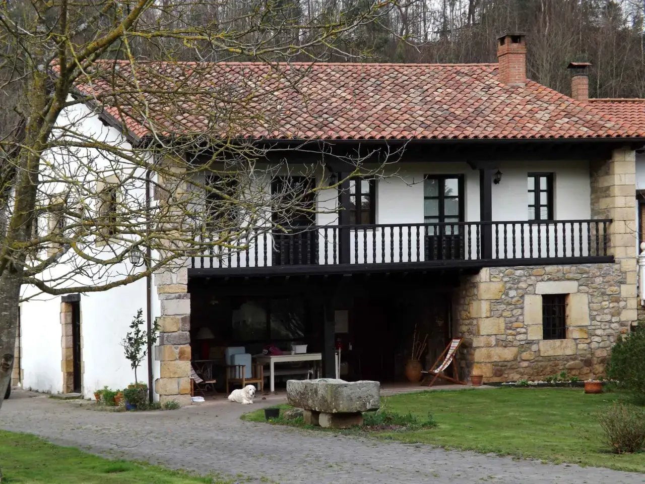 Una casa típica de Cantabria, con tejado de tejas rojas, paredes blancas y detalles de piedra. Un perro blanco descansa en el porche.