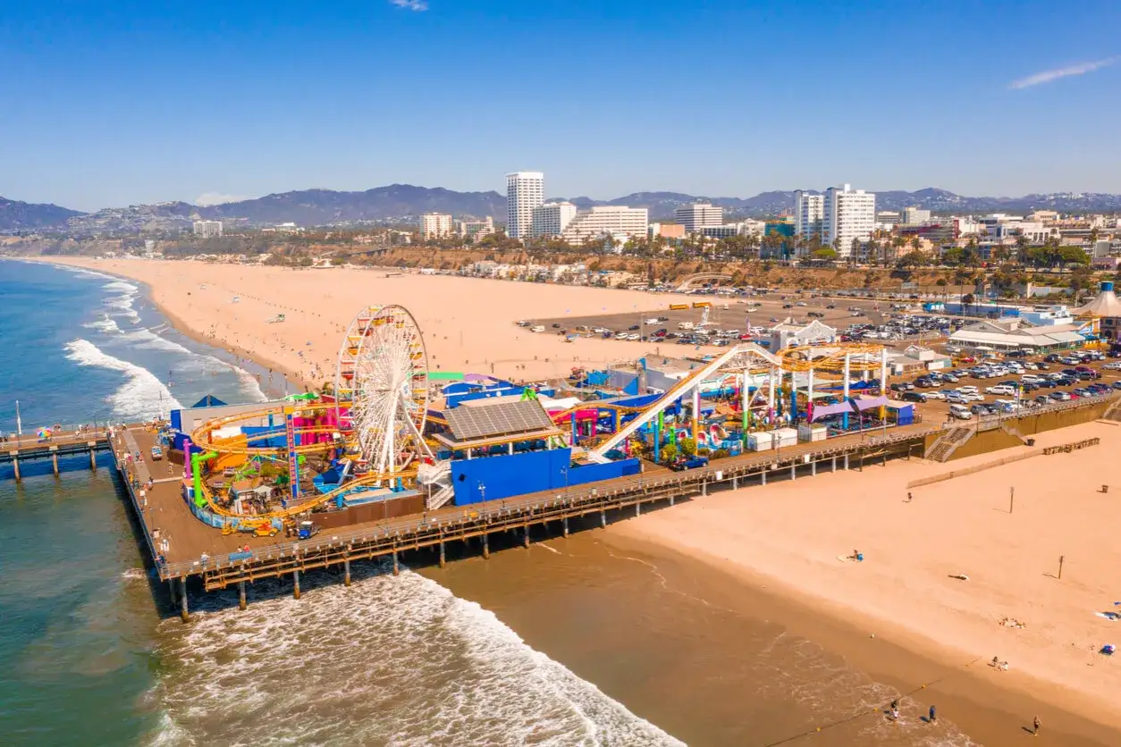 Wspaniała pogoda w Los Angeles w lutym. Molo w Santa Monica z diabelskim młynem i parkiem rozrywki, plaża pełna ludzi, ocean i góry w tle.