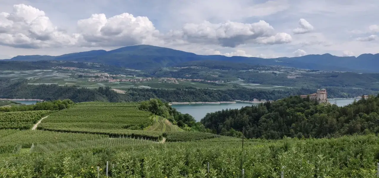 Vasta campagna con meleti rigogliosi e un lago azzurro, dominata da montagne e un castello. La forcella di Brez offre uno scenario incantevole.