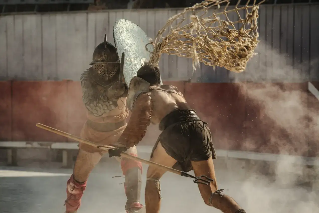 Deux gladiateurs romains s'affrontent dans l'arène, la poussière soulevée par leurs mouvements. L'un lance un filet, l'autre se protège avec son bouclier.