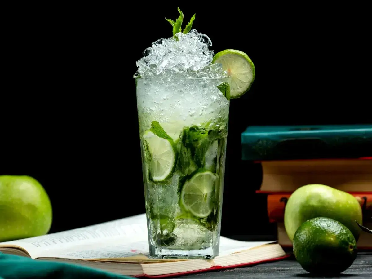 Orzeźwiający mojito z wódką, pełen mięty i limonki, podany z lodem. Obok jabłka i książki, idealny na letnie popołudnie.