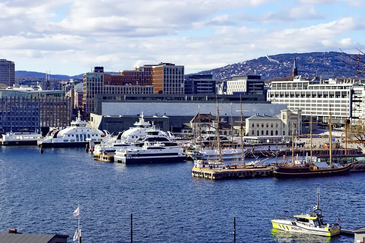 Navi moderne e velieri ormeggiati nel porto di Oslo, con edifici contemporanei e colline sullo sfondo.