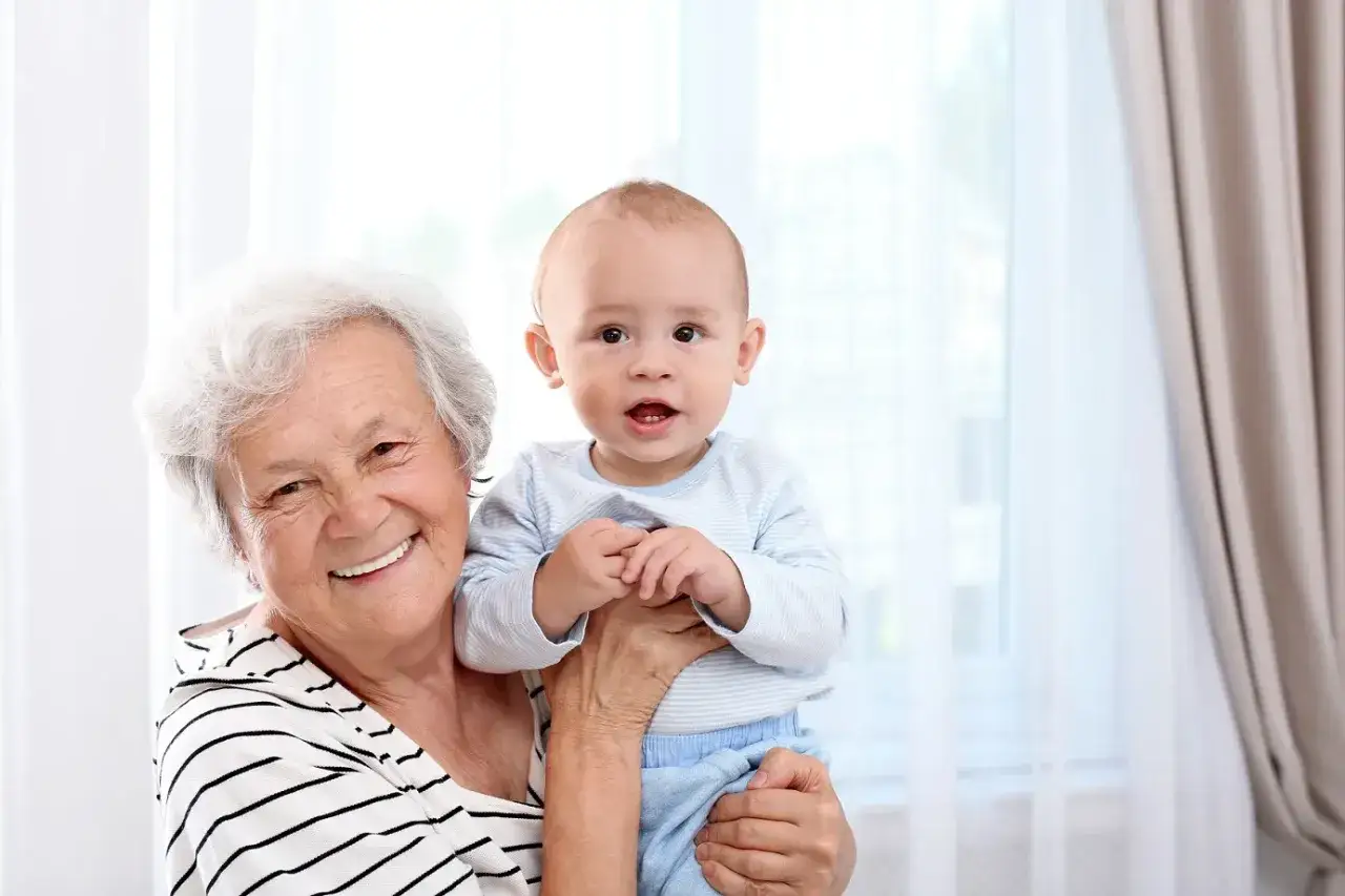 Uśmiechnięta babcia trzyma wnuka. Zastanawiasz się, ile kosztuje niania na pełen etat?