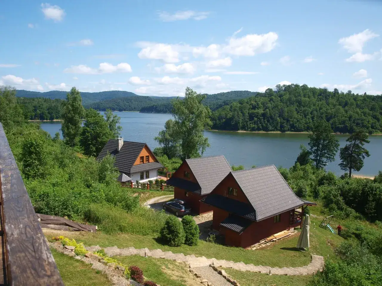 Olchowiec nad Soliną, z widokiem na jezioro i zalesione wzgórza. Dwa drewniane domki i biały budynek z czerwonym dachem.