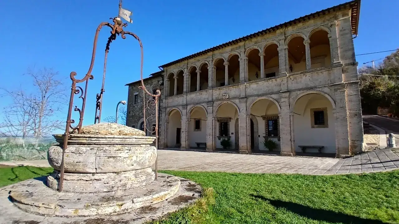 Antico pozzo in pietra davanti al tempio di San Biagio, con un portico ad archi e cielo azzurro.