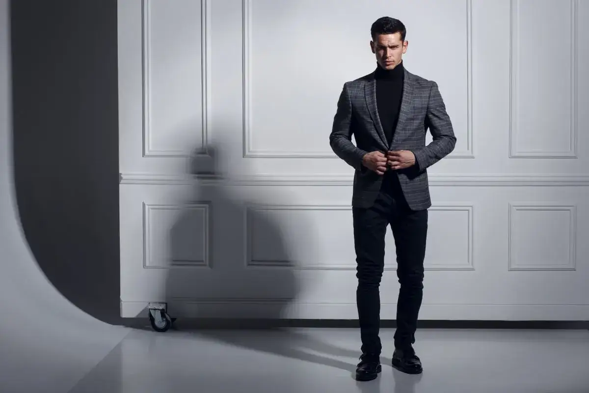 Uomo con un elegante outfit con dolcevita nero, blazer a quadri grigio e pantaloni scuri.