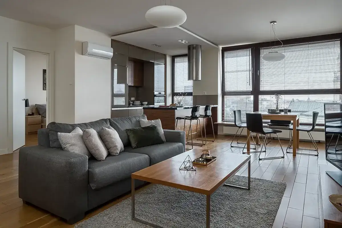 Nowoczesna klimatyzacja na 40m2 zapewnia komfort w salonie z aneksem kuchennym. Szara sofa, drewniany stolik i jadalnia tworzą przytulną przestrzeń.