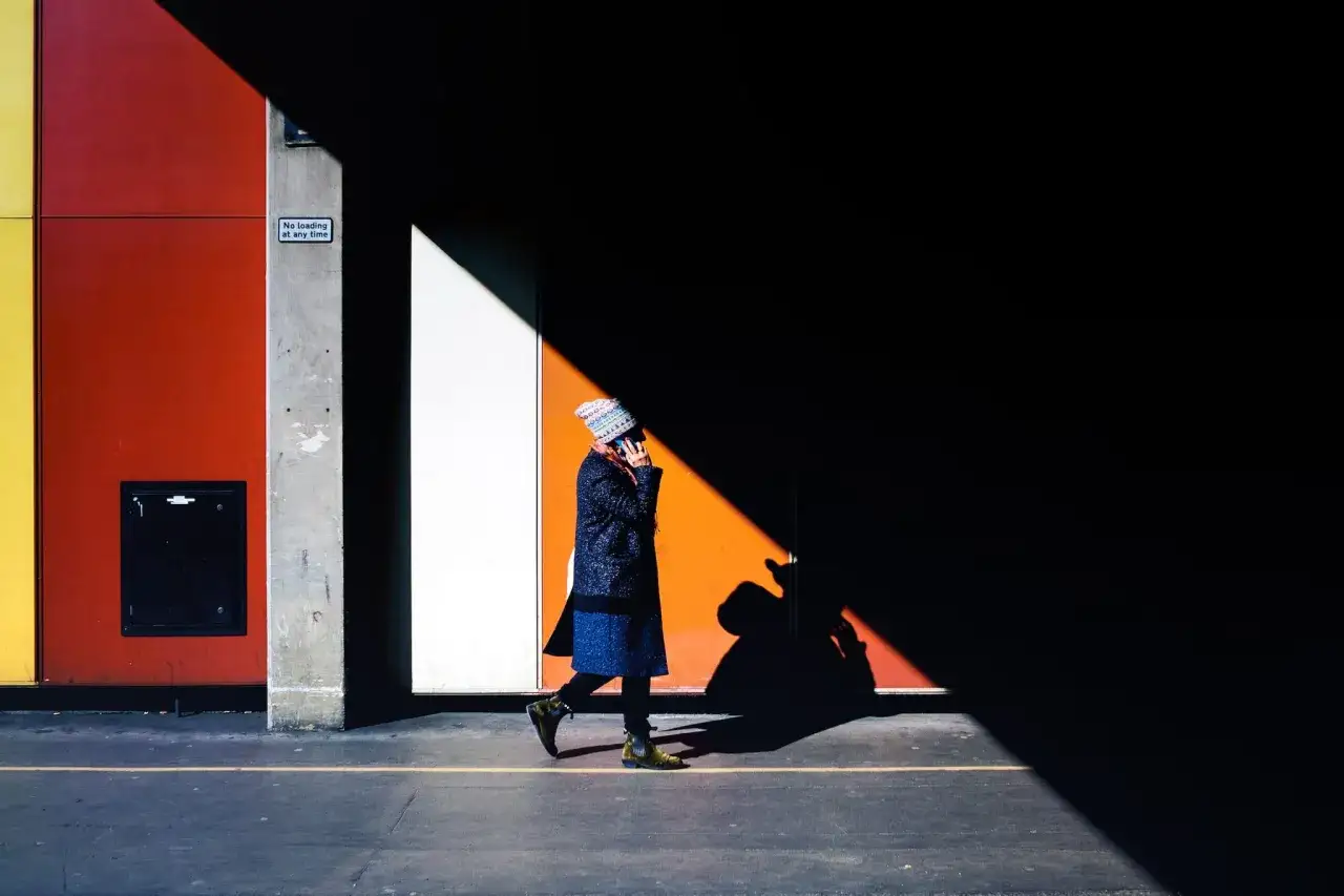 Street foto: kobieta w kolorowym kapeluszu idzie w cieniu, na tle geometrycznej ściany w odcieniach ż&oacute;łci, czerwieni i bieli.