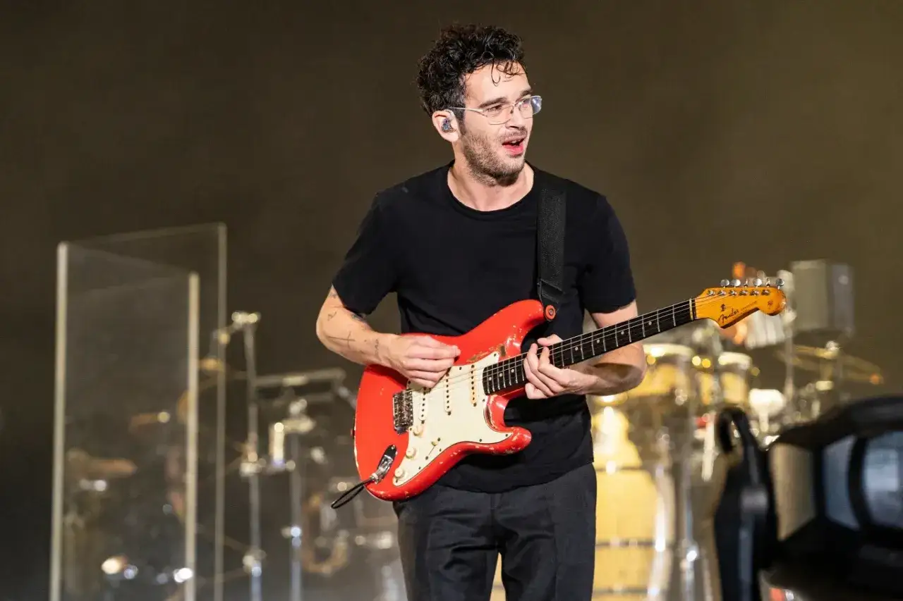 Matty Healy, w okularach i czarnej koszulce, gra na czerwonej gitarze elektrycznej na scenie.