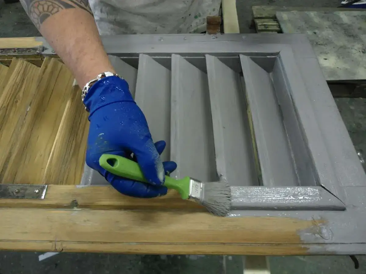 Mano con guanto blu che dipinge con pennello verde un infisso in legno, progetto fai da te per rinnovare.