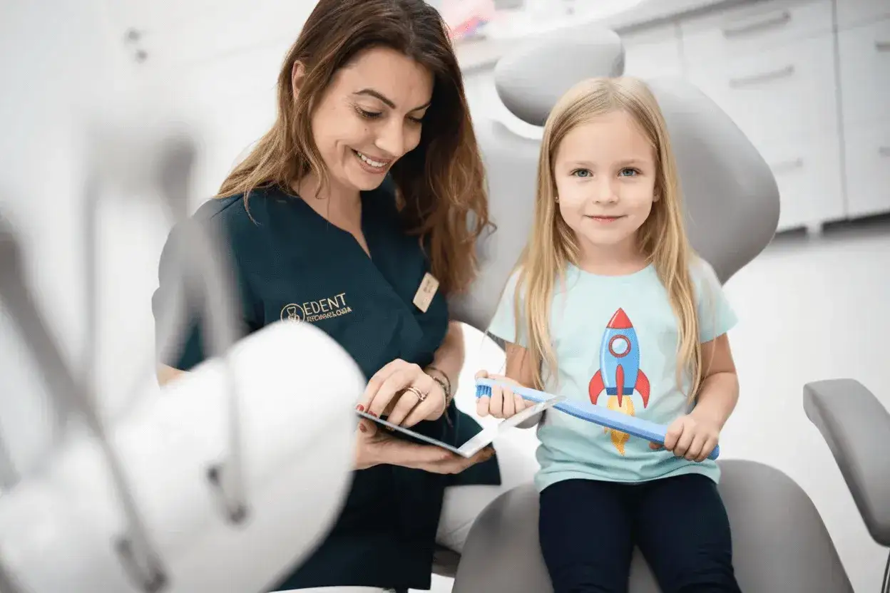 Uśmiechnięta dentystka pokazuje dziecku, jak używać szczoteczki z fluorkiem sodu, by mieć zdrowe zęby.