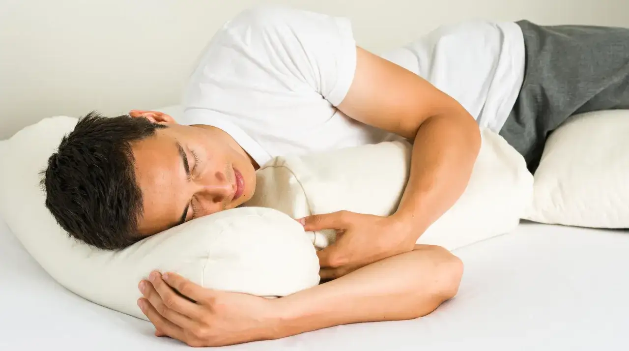 Uomo dorme sul fianco sinistro, abbracciando un cuscino. La posizione per dormire a destra o sinistra pu&ograve; influenzare il riposo.
