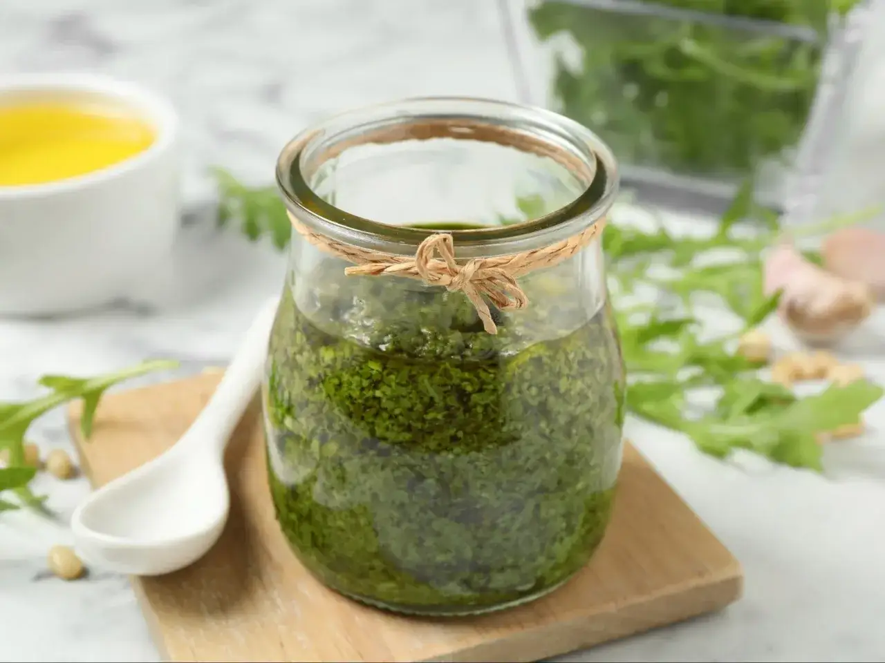 Słoik pełen aromatycznego pesto z rukoli, obok biała łyżeczka, orzeszki piniowe i świeże liście rukoli.