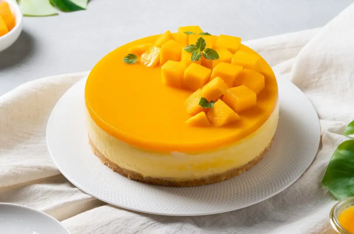 Sernik z błyszczącą polewą mango, ozdobiony kostkami owocu i listkami mięty.