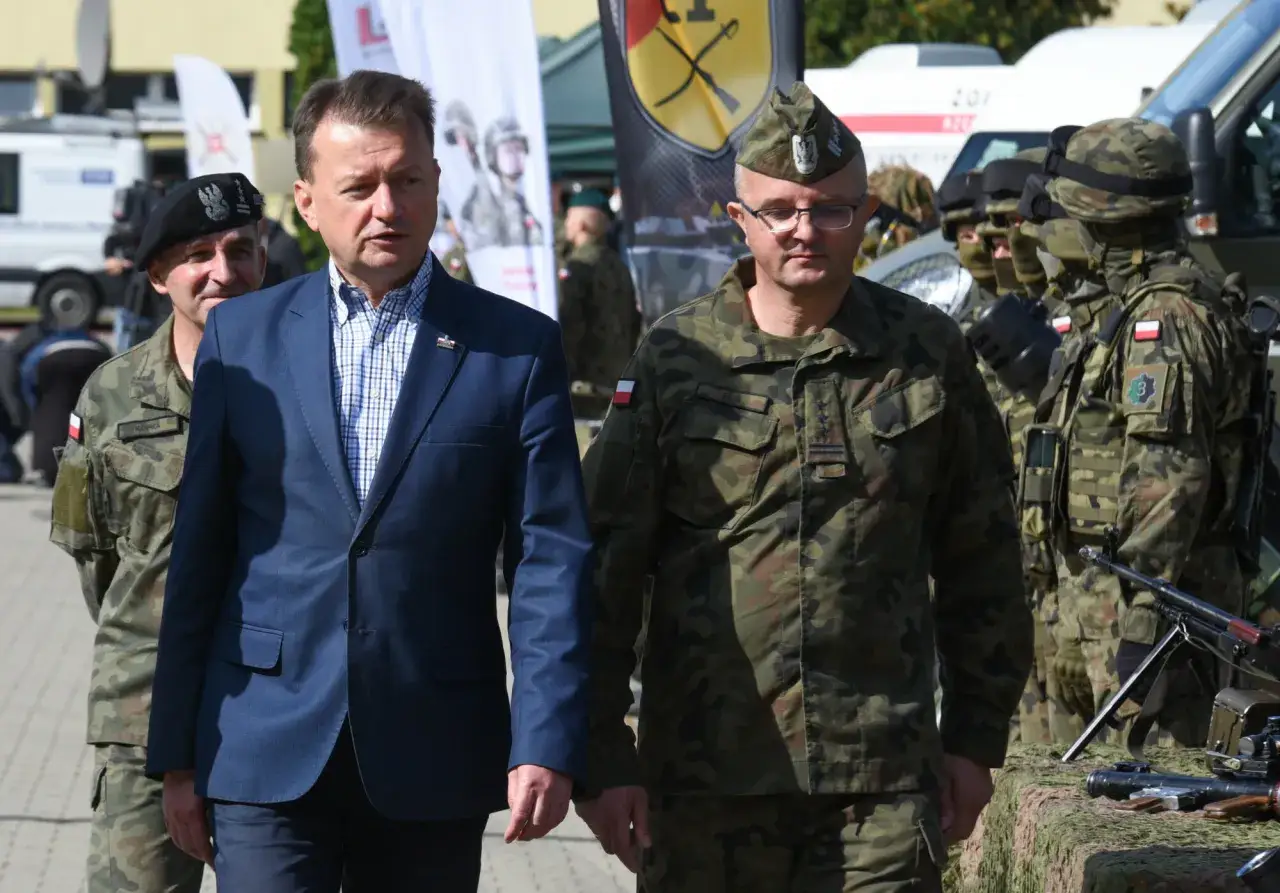 Dwóch żołnierzy w mundurach i mężczyzna w garniturze. Czy wojsko to budżetówka? W tle flagi i sprzęt wojskowy.