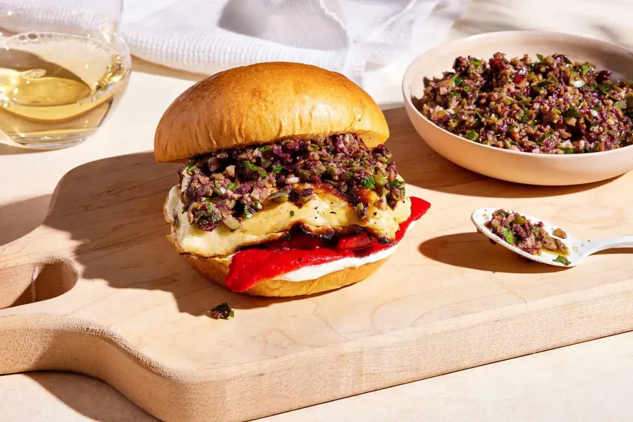 Saftiger halloumi burger mit Oliven-Topping und roten Paprika auf einem Holzbrett. Daneben eine Schüssel mit dem Topping und ein Löffel.