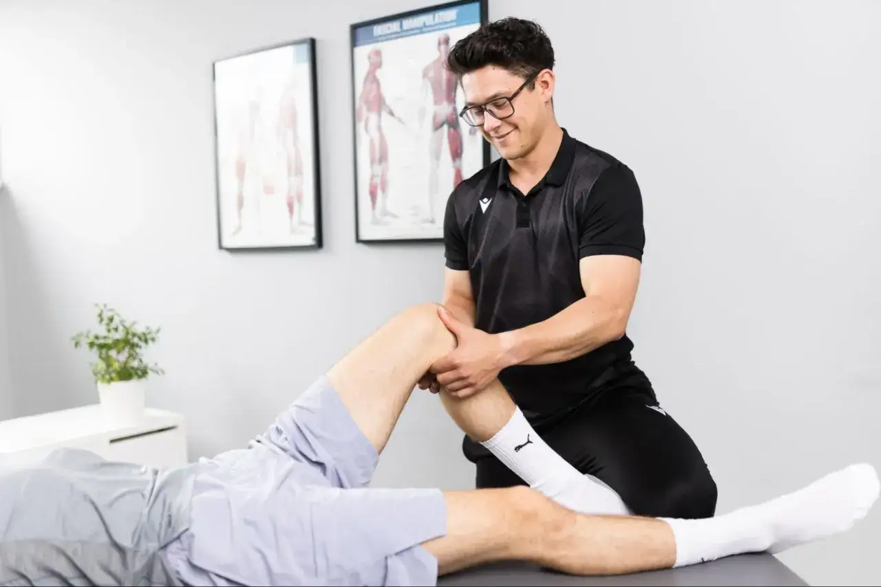 Fizjoterapeuta wykonuje masaż powięziowy kolana pacjenta. Na ścianie wiszą anatomiczne plakaty.