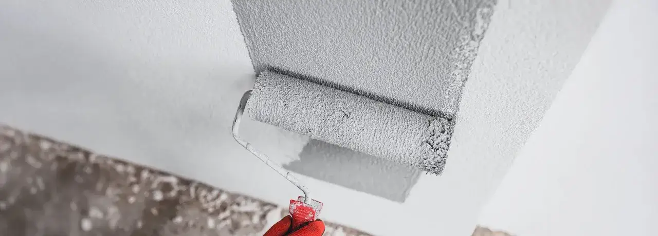 Eine rote Handschuh-tragende Hand grundiert mit einer Farbrolle eine raue Wand in Grau.