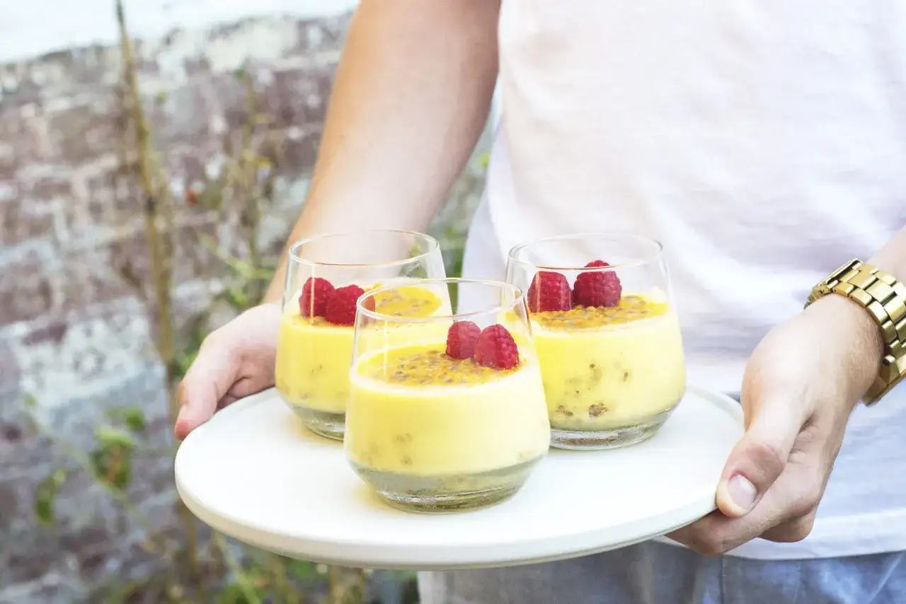 Tres copas de cremoso mousse de maracuya, decoradas con frambuesas y semillas de maracuyá, servidas en una bandeja blanca.