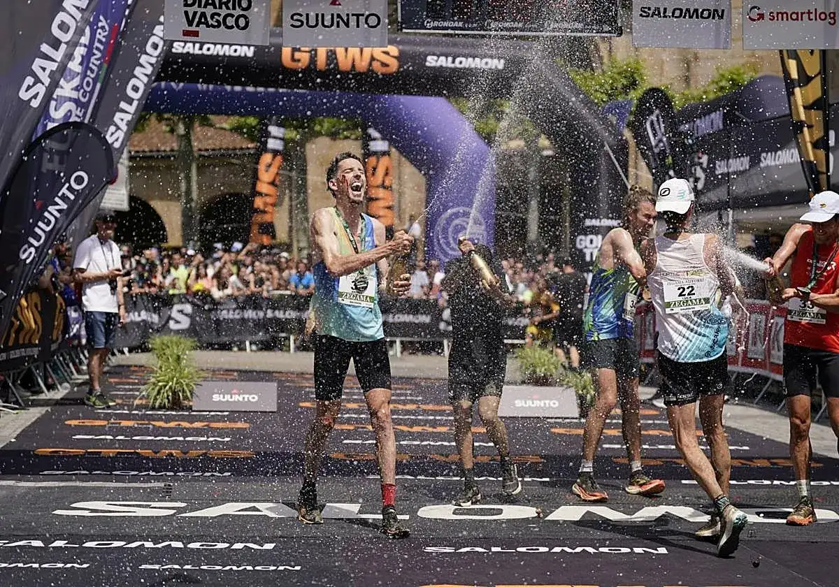 Ganadores del **Zegama-Aizkorri** celebran con champ&aacute;n. Corredores empapados de espuma bajo un arco de meta.
