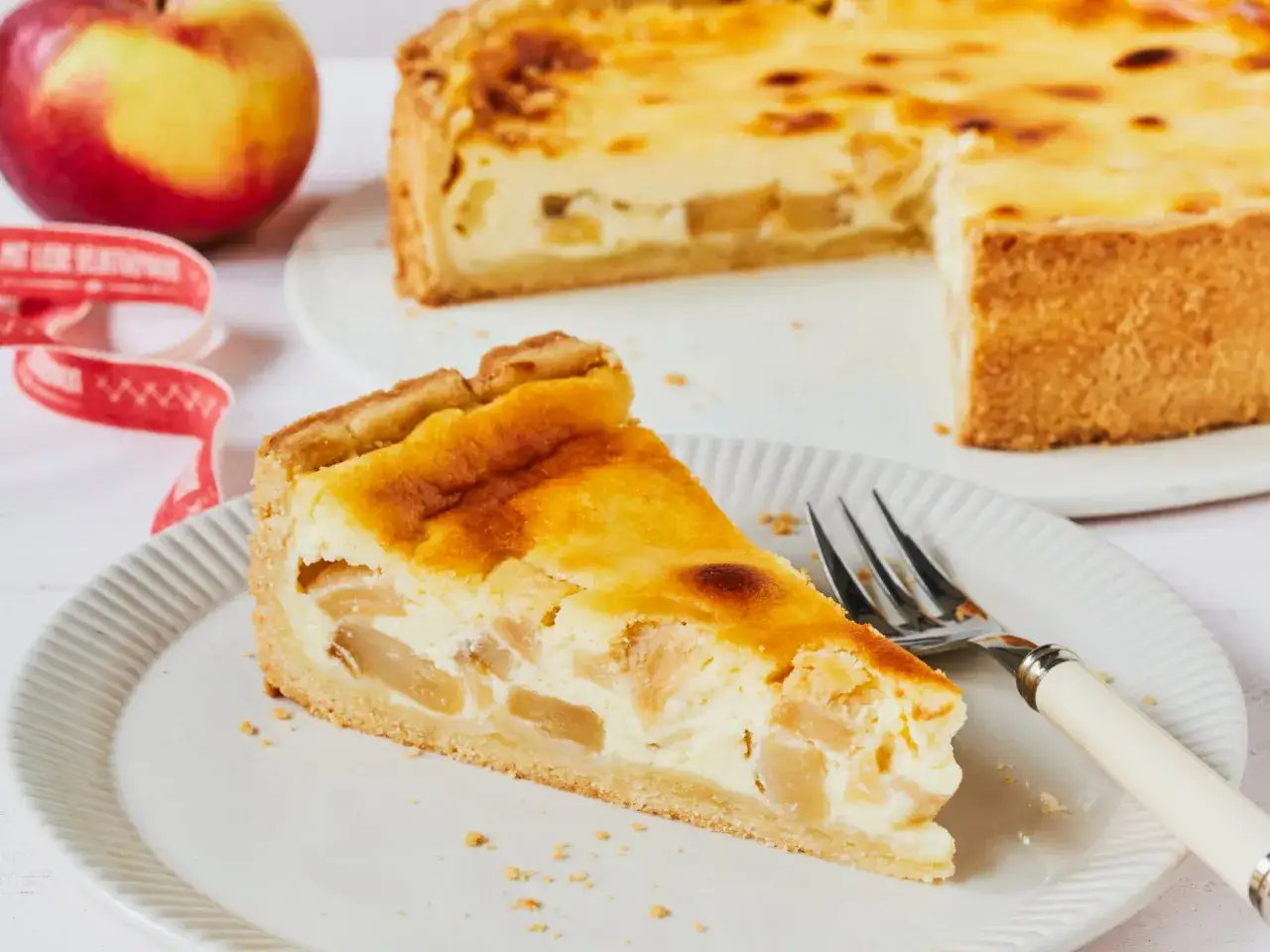 Ein St&uuml;ck saftiger Apfelkuchen mit Quark auf einem Teller, daneben ein ganzer Kuchen und ein Apfel.