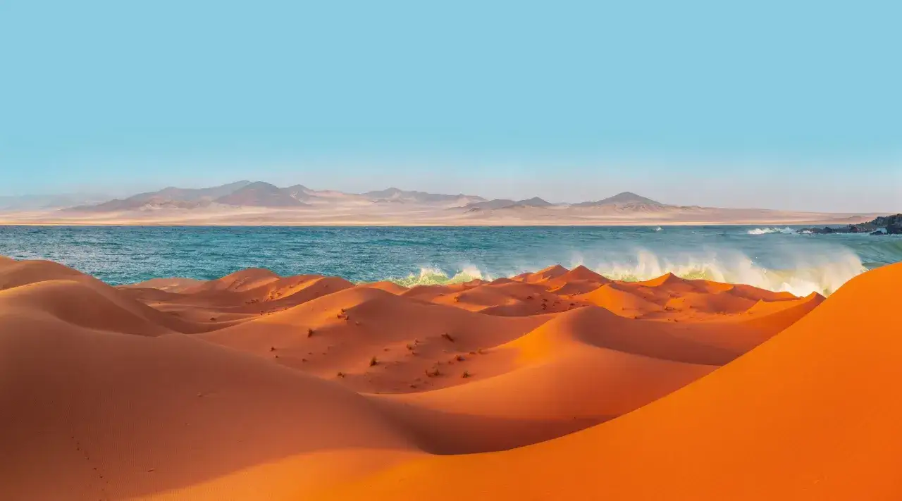 Pomarańczowe wydmy pustyni Namib spotykają się z turkusowym oceanem. W oddali widać g&oacute;ry.