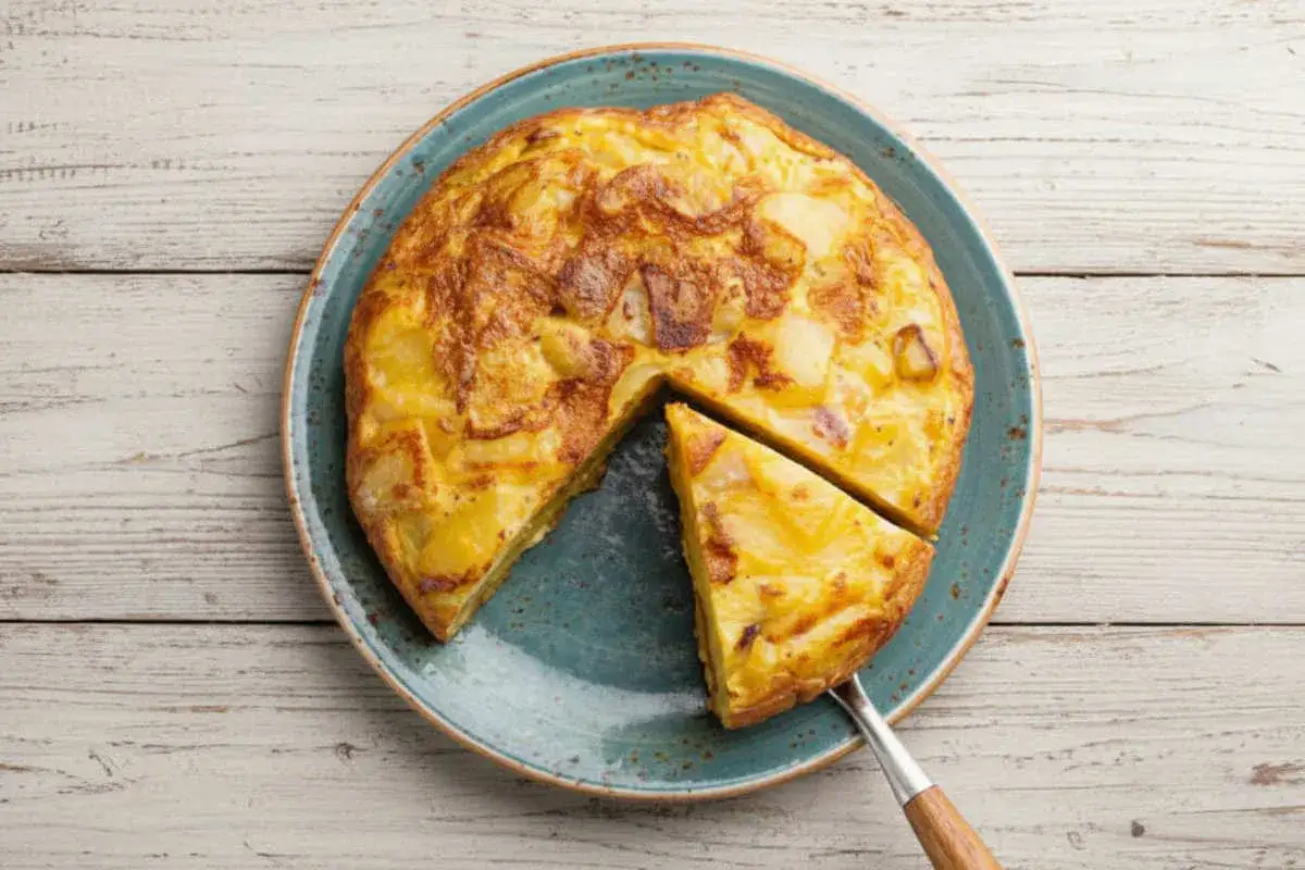 Une délicieuse tortilla espagnole, dorée et moelleuse, est servie sur une assiette bleue. Une part est déjà coupée.