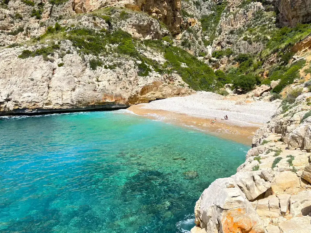 Aguas turquesas bañan la cala dels Testos, una joya escondida entre acantilados rocosos y vegetación. Dos personas disfrutan de la tranquilidad en su arena.